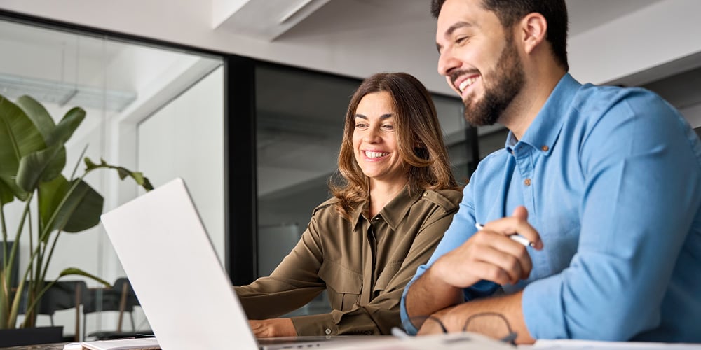 Two people seated at a desk reviewing information together on a laptop.