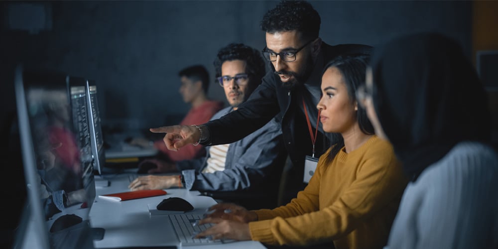 Group in a computer lab as one person points at a monitor while others follow along at workstations.