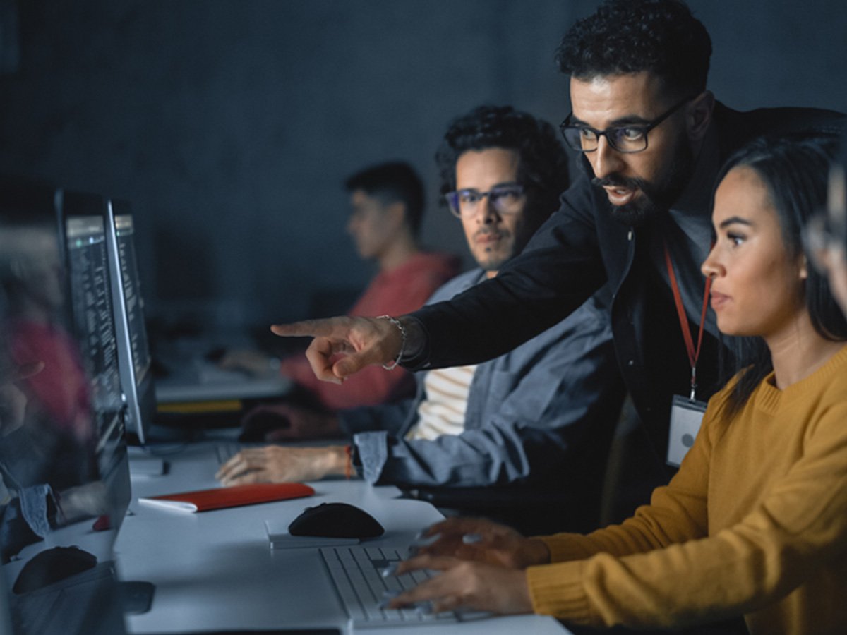 Group in a computer lab as one person points at a monitor while others follow along at workstations.