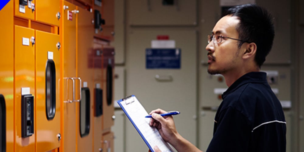 Worker checking electrical cabinets and recording details on a clipboard in an industrial facility.