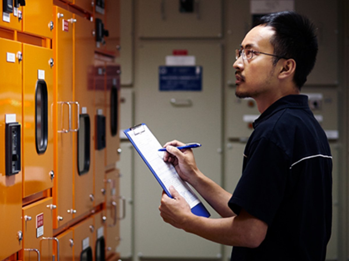 Worker checking electrical cabinets and recording details on a clipboard in an industrial facility.