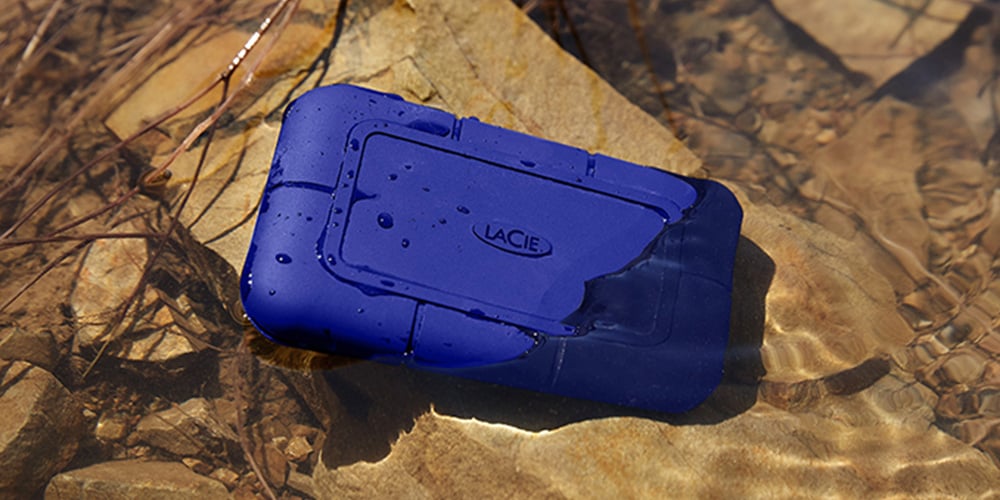 Blue LaCie rugged hard drive partially submerged in shallow water.
