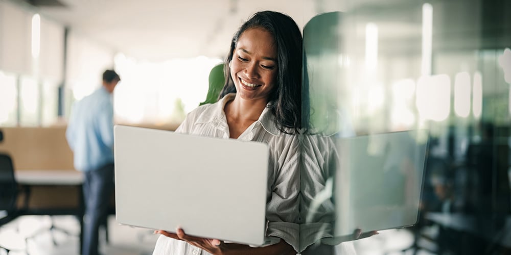 Individual standing in a modern office holding an open laptop beside a glass partition.