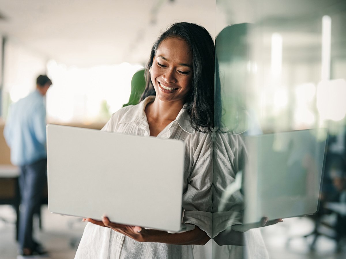 Individual standing in a modern office holding an open laptop beside a glass partition.