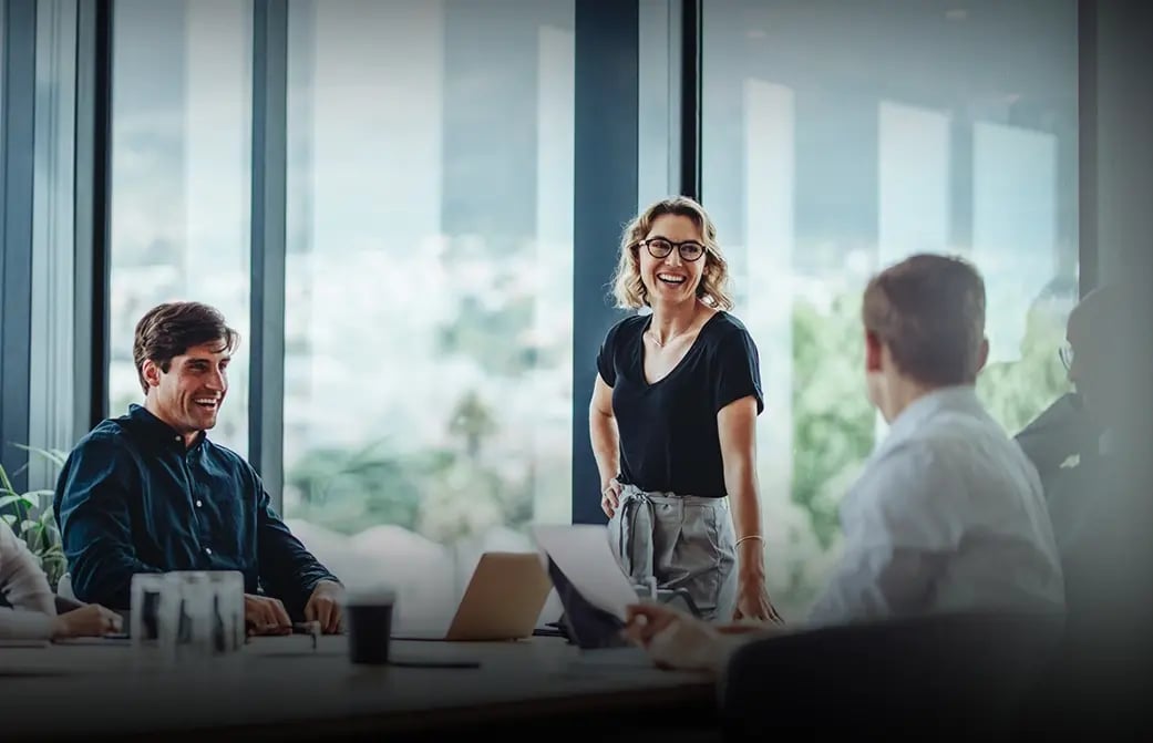 Person standing and presenting to colleagues seated around a table in a meeting room.