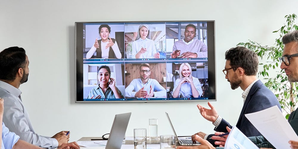 Team in a meeting room participating in a video conference displayed on a large screen.