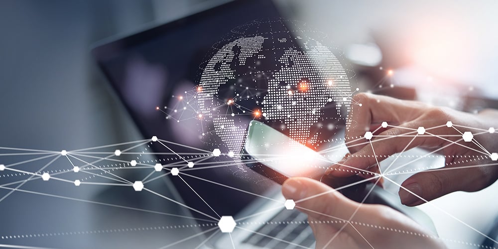 Hands using a smartphone over a laptop with a digital globe and network graphics overlay.