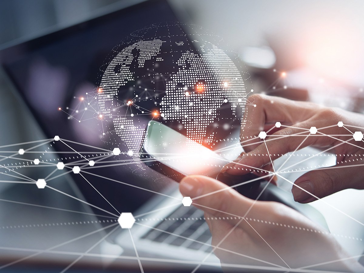 Hands using a smartphone over a laptop with a digital globe and network graphics overlay.