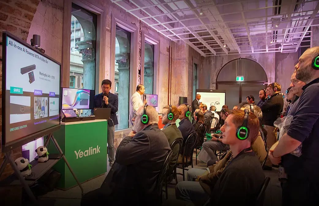 Audience wearing headsets listens to a presenter demonstrating communication technology at a large indoor event.