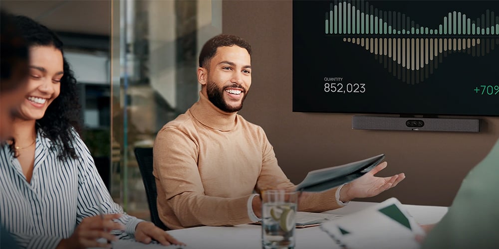 Colleagues in a meeting as one person presents data on a wall-mounted screen showing charts and growth figures.