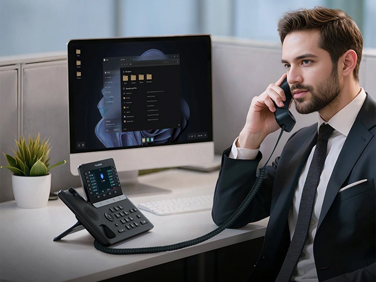 Business professional speaking on a desk phone at a modern workstation with a computer, keyboard, and office plant.