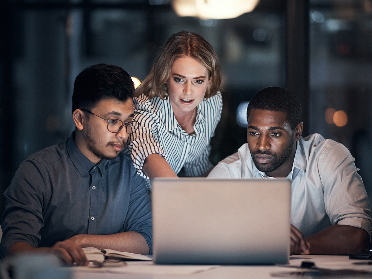 Three colleagues focus intently on a laptop in a dimly lit office, suggesting dedication and teamwork while solving a problem or project.