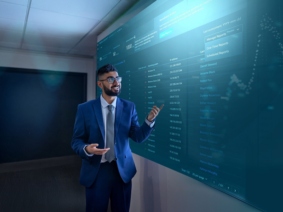 A man in a suit, smiling and gesturing toward a large digital screen displaying data and graphs. The setting is professional and tech-oriented.