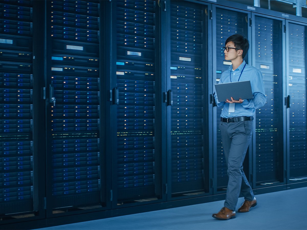 Person holding a laptop while standing in a server room with rows of illuminated server racks.