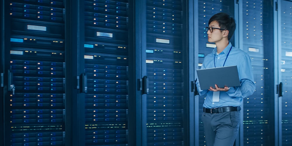 Person holding a laptop while standing in a server room with rows of illuminated server racks.