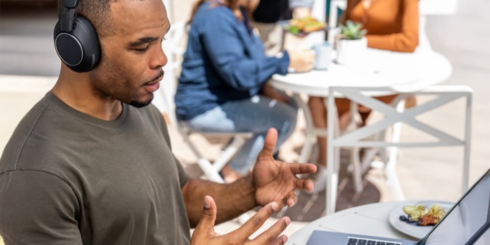 A man with headphones gestures while on a video call with a laptop at an outdoor cafe. In the background, people sit at another table.