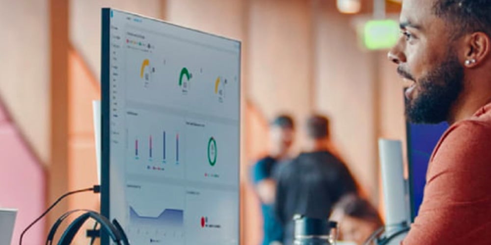 A man in a red shirt looks at a computer screen displaying graphs and charts in an office environment. The scene conveys focus and productivity.