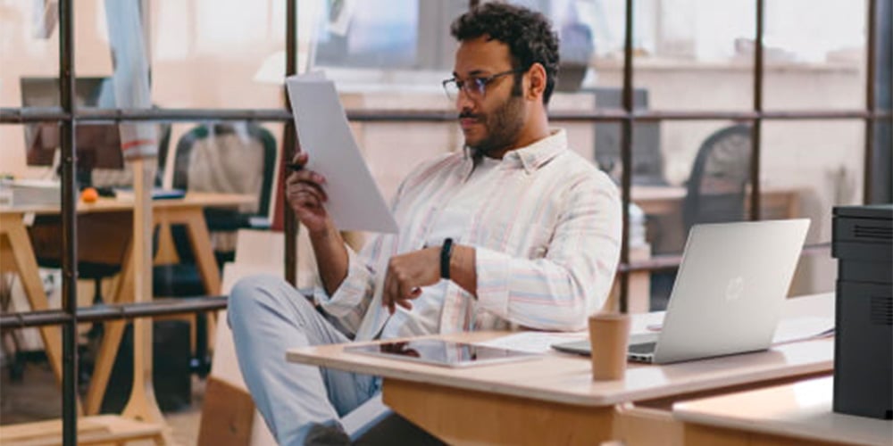 A man in a casual shirt sits at a desk in a modern office, intently reading a paper. A laptop, phone, and coffee cup are nearby, suggesting a focused work environment.