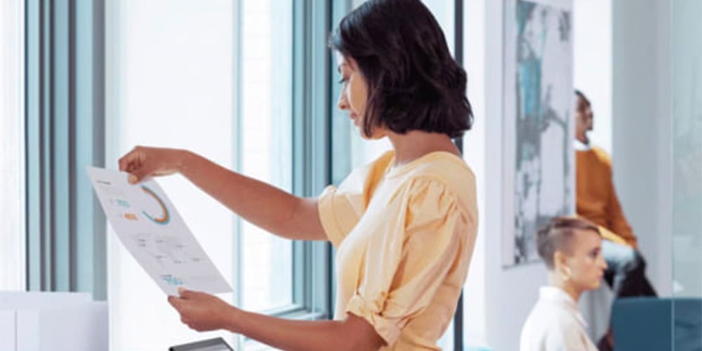 Woman in yellow blouse reviews a graph-filled document by a bright office window. Background shows two people working, adds a focused atmosphere.