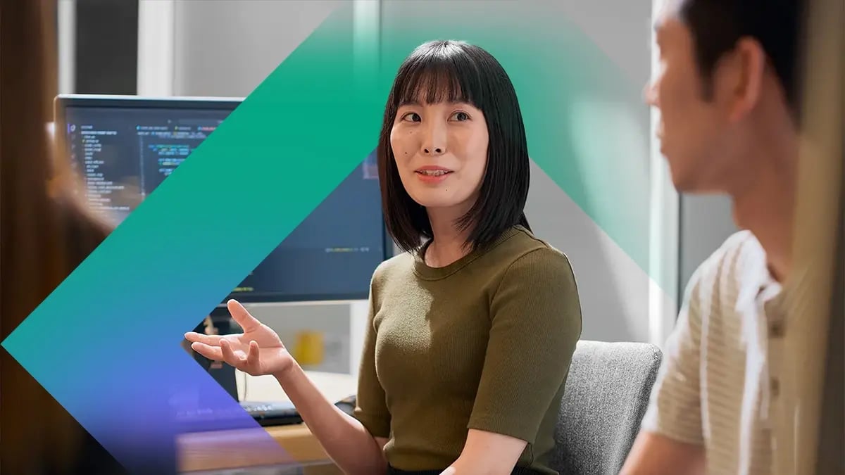 A person gestures while speaking in an office setting, with colleagues nearby and a computer monitor displaying code in the background.