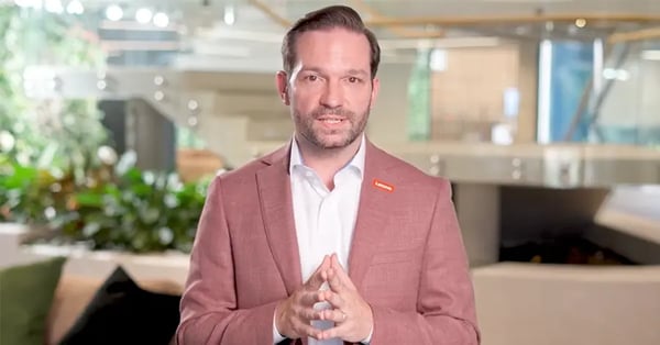 Person in a blazer standing indoors, speaking to camera in a modern office setting with plants in the background.