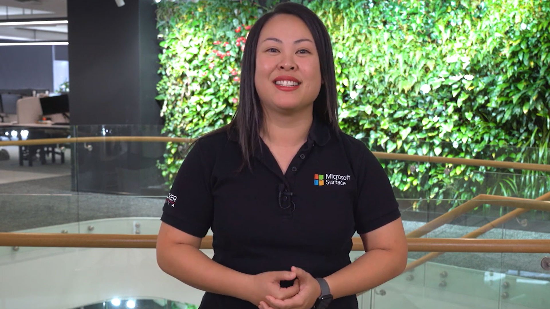 Smiling woman in a black Microsoft Surface shirt stands in front of a lush, green vertical garden wall, with a modern office setting in the background.