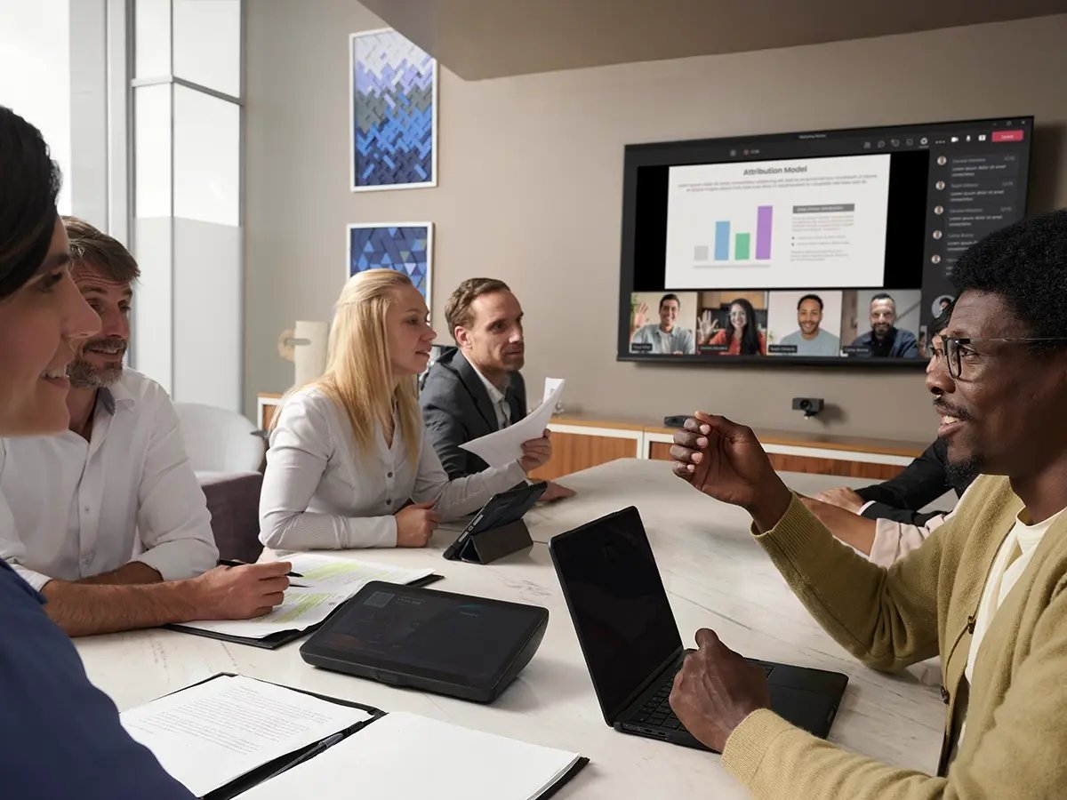 A group of people in a conference room participating in a hybrid meeting, with a presentation and video call displayed on a large screen.