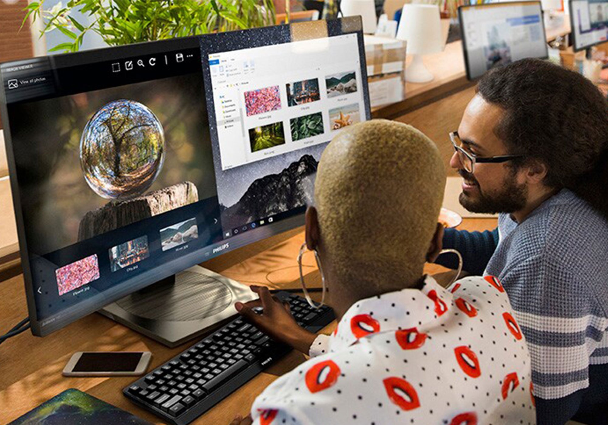 Two people working at a desk, editing images on large computer monitors in a creative workspace.